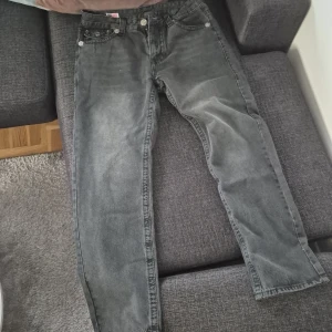Svarta jeans från True Religion - Säljer ett par svarta True Religion jeans med raka ben och klassiska detaljerade bakfickor. Jeansen har silverfärgade knappar och en tydlig logga på insidan. Tillverkade i USA och passar dig som gillar streetwear.