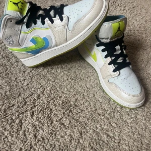  Air Jordans 1 Mid SE Warped Swoosh - Säljer ett par Air Jordans 1 Mid med vit bas, svarta skosnören och coola neon-gula och blå detaljer på sidan. Sulan är i en stark gul färg som verkligen sticker ut. Perfekta sneakers för dig som vill ha något unikt och färgglatt. Kommer UTAN originella boxen!