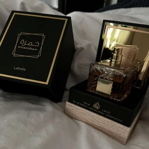 Khamrah Eau de Parfum från Lattafa - Elegant parfymflaska med guldiga detaljer och en lyxig förpackning. Khamrah från Lattafa erbjuder en sofistikerad doftupplevelse. Flaskan har en fyrkantig form med ett dekorativt mönster och kommer i en stilren svart och guldig kartong.