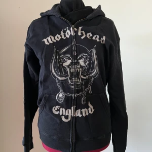Svart Motörhead hoodie med dragkedja - Säljer en svart hoodie med dragkedja från Motörhead. Framsidan har ett stort tryck med bandets logga och texten 'Motörhead England'. Baksidan pryds av texten 'Everything Louder Than Everything Else'. 