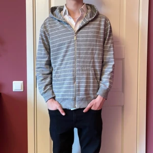 Grå randig hoodie med dragkedja - Säljer en stilren grå hoodie med vita ränder och dragkedja framtill. Tröjan har en bekväm passform med långa ärmar och en praktisk huva. Perfekt för en avslappnad look.