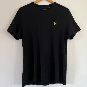 Svart t-shirt med gul logga - Säljer en stilren svart t-shirt från Lyle & Scott med en liten gul logga på bröstet. T-shirten har en klassisk passform och är tillverkad i mjuk bomull. 