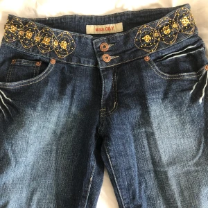Blå jeans med broderade detaljer från Miss D&Y - Snygga blå jeans med unika broderade detaljer i guld och svart på midjan och bakfickorna. Jeansen har en klassisk femficksdesign och knappar framtill. Perfekta för att ge din outfit en extra touch.