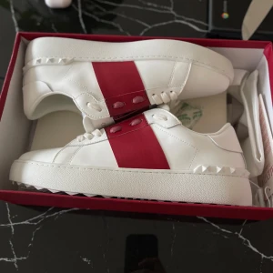 Valentino opens - Snygga vita sneakers från Valentino med en bred röd rem över ovansidan. Skorna har en rund tå och dekorativa nitar längs sulan. Perfekta för en stilren look. Säljer pga stor storlek 