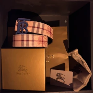 Rutig Burberry bälte med silverfärgat spänne - Säljer ett snyggt Burberry bälte med klassiskt rutig design i beige, brun och röd. Bältet har ett stort silverfärgat 'TB'-spänne och är tillverkat i skinn med tygöverdrag. Perfekt accessoar för att lyfta din outfit.