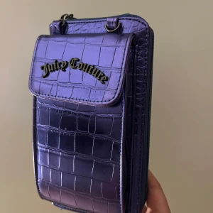 Lila axelväska från Juicy Couture - Säljer en lila axelväska från Juicy Couture i glansigt syntetmaterial med krokodilpräglad yta. Väskan har flera fack med dragkedja och ett lock framtill med metallogo. Perfekt för mobilen och småsaker.