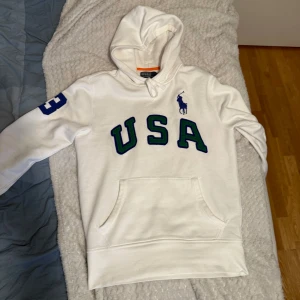 Vit hoodie från Polo Ralph Lauren med USA-tryck - Vit hoodie från Polo Ralph Lauren med stor USA-text i blått och grönt framtill, blå siffra 3 på ärmen och klassisk broderad logga på bröstet. Mjuk bomullskvalitet och känguruficka. Inga fläckar och i väldigt fint skick! Va inte rädd för ställa frågor!