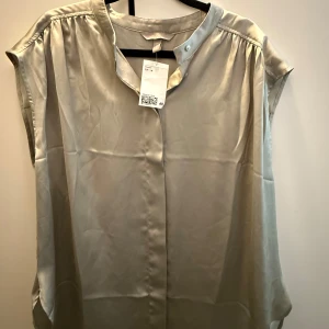 Blus från H&M - Säljer en stilren grön ärmlös blus från H&M. Blusen har en mjuk, glansig yta och knäppning vid halsen. Perfekt för en enkel och elegant look. Blusen är mintgrön i verkligheten vilket framgår mer på den andra bilden. Aldrig använd, helt ny med prislapp💚