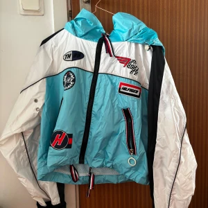 TOMMY HILFIGER vindjacka Gigi Hadid - Säljer en sportig turkos och vit vindjacka med svarta detaljer och flera coola patchar framtill. Jackan har huva, dragkedja och snörning i nederkant. Perfekt för dig som gillar en färgstark och unik stil.