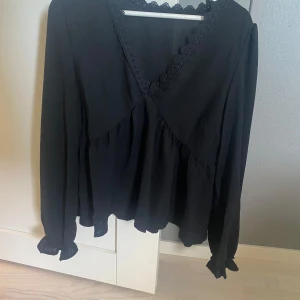 Svart blus med spetsdetaljer - Snygg svart blus med v-ringning och spetsdetaljer runt halsen. Blusen har långa ärmar och en lös passform som ger en elegant look. Perfekt för både vardag och festliga tillfällen.