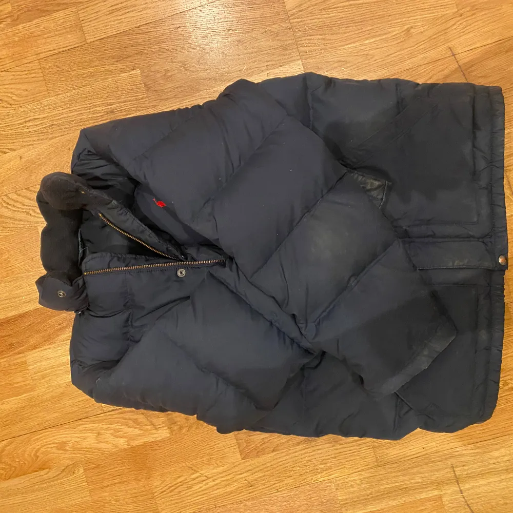 Säljer en mörkblå dunjacka från Polo Ralph Lauren med klassisk röd logga på bröstet. Jackan har hög krage, tryckknappar och dragkedja samt stora fickor framtill. Perfekt för kalla dagar och har en tidlös look.. Takit.