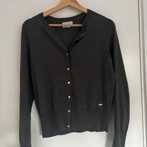 Mörkgrå kofta från Filippa K - Stilren mörkgrå kofta från Filippa K med långa ärmar och knappar framtill. Klassisk design med ribbade muddar och diskret metallogo vid nederkanten. Perfekt för lager-på-lager och enkel att matcha.