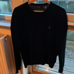 Svart merinoullströja från Polo Ralph Lauren - Säljer en klassisk svart tröja från Polo Ralph Lauren i mjuk merinoull. Tröjan har rund halsringning, lång ärm och den ikoniska broderade loggan på bröstet. Perfekt för lager-på-lager och stilrena outfits. Hör av dig vid funderingar!