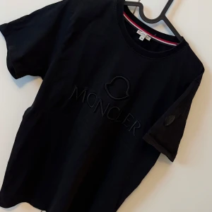 Svart t-shirt från Moncler - Säljer en svart t-shirt från Moncler med diskret broderad logga på bröstet och ärmen. Klassisk rund halsringning och korta ärmar. Perfekt för dig som gillar stilrena märkesplagg.