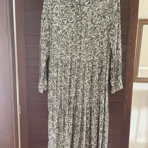 Grön blommig långklänning från H&M - Säljer en långklänning från H&M i grön nyans med vitt blommönster. Klänningen har lång ärm, klassisk krage och knäppning framtill. Perfekt för dig som gillar en avslappnad men ändå stilren look.