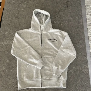Grå hoodie med tryck från Zara - Grå hoodie från Zara med svart tryck 'Oakland Park San Jose Road' på både bröstet och ryggen. Tröjan har dragkedja framtill, huva och två fickor. Perfekt för en avslappnad stil.