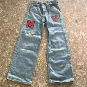 Ljusblå wide jeans med patches - Ljusblå jeans med breda ben och hög midja från H&M. Byxorna har coola röda patches, bland annat ett stort M och en gitarr på framsidan. Justerbar midja för extra komfort. Perfekta för en avslappnad och trendig stil.