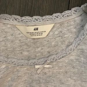 Grå långärmad topp från H&M - Säljer en söt grå långärmad topp från H&M med spetsdetaljer vid halsringningen och en liten rosett framtill. Perfekt för en stilren och bekväm look. 💫