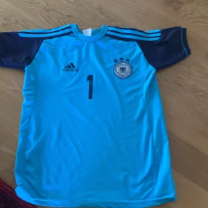 Manuel Neuer tröja Tyskland - Säljer en blå fotbollströja från Adidas med korta ärmar. Tröjan har ett tryck med nummer 1 och texten 'Neuer' på baksidan. Den har även ett emblem för det tyska fotbollsförbundet på framsidan. Den är nästan oanvänd och köpt i Tyskland.
