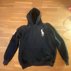 Svart hoodie från Polo Ralph Lauren - Säljer en svart hoodie från Polo Ralph Lauren i klassisk passform. Tröjan har en stor vit broderad logga på bröstet, huva och magficka. Perfekt för dig som gillar stilrena och tidlösa plagg.