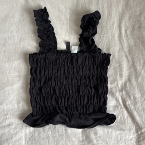 Svart smockad linnetopp från HM - Säljer en svart smockad linnetopp från HM med breda volangprydda axelband och rynkad struktur. Toppen har en lätt volang nertill och är perfekt för varma dagar.