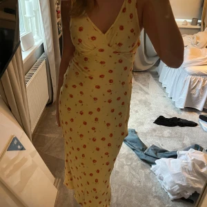 Gul blommig klänning - Säljer en söt gul klänning med blommigt mönster. Klänningen har en v-ringad design och är ärmlös, perfekt för varma dagar. Den är lång och har en smickrande passform. Använd en gång