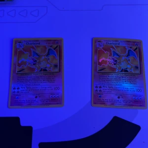 Charizard Pokémonkort - Två Charizard Pokémonkort med 120 HP. Kortens design har en bild av Charizard med eldflammor i bakgrunden. Texten beskriver Pokémon Power: Energy Burn och attacken Fire Spin. Perfekt för samlare eller Pokémon-entusiaster!