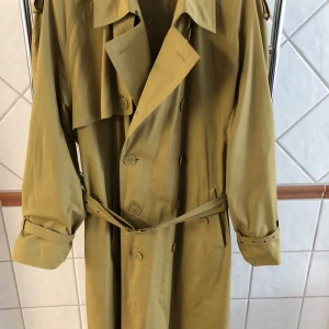 Grönbeige trenchcoat från Yves Saint Laurent - Klassisk grönbeige trenchcoat från Yves Saint Laurent med knappar och bälte i midjan. Jackan har långa ärmar, bred krage och axelklaffar. Perfekt för dig som vill ha en tidlös och stilren look.