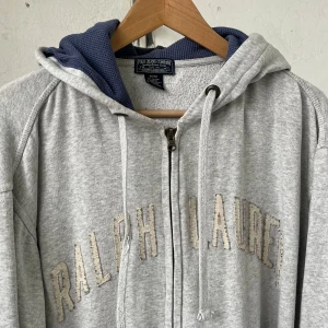 Grå hoodie från Ralph Lauren - Grå hoodie från Ralph Lauren med dragkedja och broderad logga framtill. Jackan har huva med blått innerfoder och justerbara snören. Perfekt för en avslappnad stil. Är storlek m, men passar också large. Den är vintage, och får en se ut som en grabb från nån amerikansk college film som kör amerikansk fotboll! Man ser stadig ut i den helt enkelt!