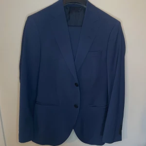 Blå kostym från Tiger of Sweden - Snygg blå kostym från Tiger of Sweden. Kavajen har två knappar och klassiska slag. Byxorna är stilrena med en knappdetalj vid bakfickan. Perfekt kostym för studenten.