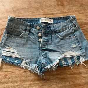 Blå jeansshorts med fransar från Abercrombie & Fitch - Snygga blå jeansshorts från Abercrombie & Fitch med låg midja, slitna detaljer och fransig kant. Knappar framtill och klassiska fickor både fram och bak. Perfekta för en avslappnad stil.