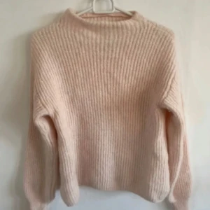 Ljusrosa stickad tröja - Mysig ljusrosa stickad tröja med ribbad struktur och hög hals. Tröjan har långa ärmar och är tillverkad av en mjuk ullblandning med alpacka, vilket gör den extra varm och skön.