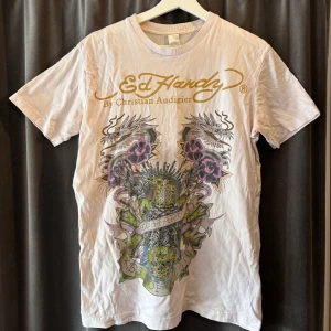 Vit Ed Hardy t-shirt - Säljer en vit/krämfärgad t-shirt från Ed Hardy med stort tryck framtill som visar två drakar, lila blommor och texten 'Kiss of Death'. T-shirten har korta ärmar och klassisk passform. Perfekt för dig som gillar street style och coola motiv.⭐️