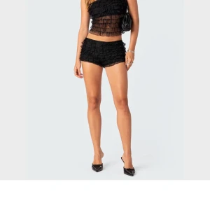 Svarta spetsiga volangshorts  - Supersnygga svarta shorts från Edikted med flera lager volanger och spetsdetaljer. De är tighta och har en feminin look, perfekta för att sticka ut. Materialet är syntet med spets och volang för extra effekt.
