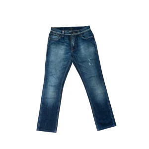Blå jeans från Nudie Jeans 29/32 - Säljer ett par klassiska blå jeans från Nudie Jeans med raka ben och snygga slitningar framtill. Jeansen har fem fickor, orangea sömmar och normal passform. Perfekta för dig som gillar en tidlös denimlook. Storleken är 29/32. Modellen är Tape Ted. 100% bomull.