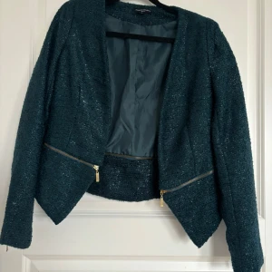 Mörkgrön kavaj med dragkedjedetaljer - Säljer en mörkgrön kavaj  med glittrig yta och guldfärgade dragkedjor framtill. Jackan har lång ärm och en snygg, asymmetrisk skärning nedtill. Perfekt för dig som vill ha en stilren men ändå unik look.