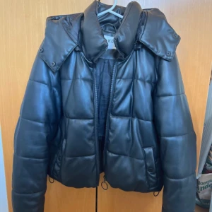 Svart  pufferjacka - Snygg svart pufferjacka med quiltad design och dragkedja. Jackan har en hög krage och avtagbar huva, perfekt för kyliga dagar. Den är både stilren och funktionell.  Det finns lite fläckar (finns bild) samt att lite av materialet på jackan slitits av.  Ingen av fickorna går att stänga då knapparna gått sönder.  Sann i strl💕