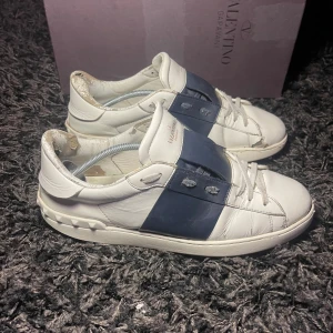 Valentino sneakers i vitt och blått - Snygga Valentino sneakers i vitt skinn med en bred blå rem över snörningen. Skorna har en stilren design med diskreta detaljer och en bekväm passform. Perfekta för att ge din outfit en lyxig touch.