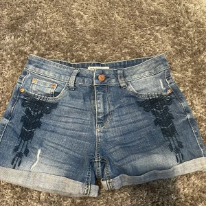 Jeansshorts med broderi - Snygga blå jeansshorts från Dr. Denim med svart broderad detalj på framsidan. Klassisk femficksmodell med uppvikta benslut och knappgylf. Perfekta för sommardagar och enkel att matcha med olika toppar.
