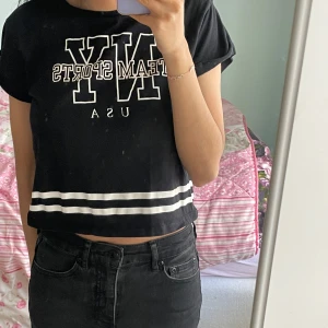 Svart croppad t-shirt från H&M med tryck - Svart croppad t-shirt från H&M med vitt tryck 'NY TEAM SPORTS USA' och två vita ränder nedtill. T-shirten har korta ärmar och en avslappnad passform. Perfekt till vardags för en sportig look.