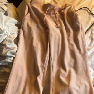 Rosa mjukisbyxor från Juicy Couture med strassdetaljer - Säljer ett par klassiska ljusrosa mjukisbyxor från Juicy Couture med strassdetaljer och logga bak. Byxorna har dragsko i midjan och raka ben. Perfekta för en chill och trendig look.