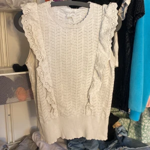 Vit ärmlös topp från Holly & Whyte - Säljer en söt vit ärmlös topp från Holly & Whyte med vackra spetsdetaljer och volanger längs axlarna. Toppen har ett stickat mönster och en rund halsringning. Perfekt för en somrig kväll med tjejerna!