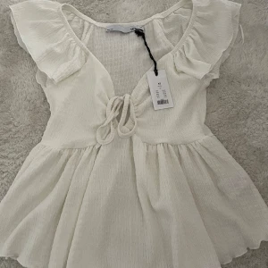 Vit blus med volangdetaljer - Säljer en söt vit blus med volangdetaljer och knytning framtill. Blusen har en luftig passform och korta ärmar med volanger. 