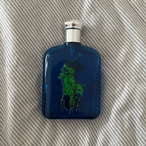 Blå parfymflaska med grön logotyp - Snygg blå parfymflaska från Ralph Lauren med en grön polo-spelare på framsidan. Flaskan har en elegant silverfärgad kork och en stilren design som passar perfekt i badrumshyllan. Riktigt billigt pris