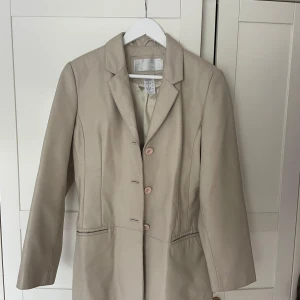 Beige skinnkavaj från La Boutique - Beige kavaj från La Boutique. Klassisk modell med krage, tre knappar framtill och två fickor.