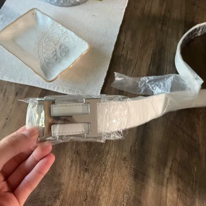 Vitt skärp med silverfärgat H-spänne - Stilrent vitt bälte med ett stort, silverfärgat H-format spänne. Bältet har en slät yta och är tillverkat i skinn. Perfekt accessoar för att lyfta din outfit med en clean och modern look.