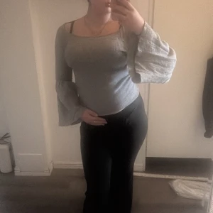 Grå ribbad topp med trumpetärmar - Supersnygg grå ribbad topp med vida trumpetärmar och figurnära passform. Toppen har en bred, något fyrkantig ringning och är tillverkad i ett stretchigt material som sitter skönt på kroppen. Perfekt för dig som gillar trendiga detaljer och vill sticka ut lite extra.