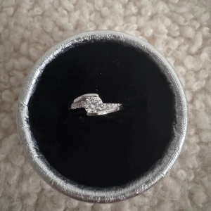 Ny ring med glittrande sten - Snygg silverfärgad ring med en stor glittrande sten i mitten och mindre stenar längs bandet. Ringen har en modern, böjd design som ger en cool och elegant vibe. Perfekt accessoar för att lyfta din stil.