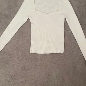 Vit ribbad långärmad topp - Snygg vit ribbad topp med lång ärm och markerad sweetheart-urringning. Figurnära passform och stretchigt material som sitter skönt. Perfekt att styla med jeans eller kjol för en clean look.