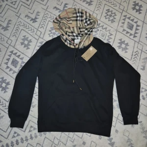 Svart hoodie från Burberry med rutmönstrad huva - Svart hoodie från Burberry med klassisk beige, svart och röd rutmönstrad huva. Tröjan har dragsko vid huvan och ribbade muddar vid ärmslut och nederkant. Perfekt för en stilren och trendig look. Har inte använt en enda gång. Lappen och allt är kvar och ser helt ny ut.